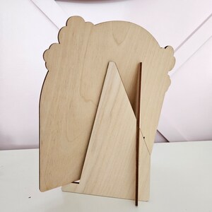 May include: A wooden picture frame with a decorative scalloped top. The frame is supported by a triangular stand and a thin wooden support. The frame is made of light-colored wood.