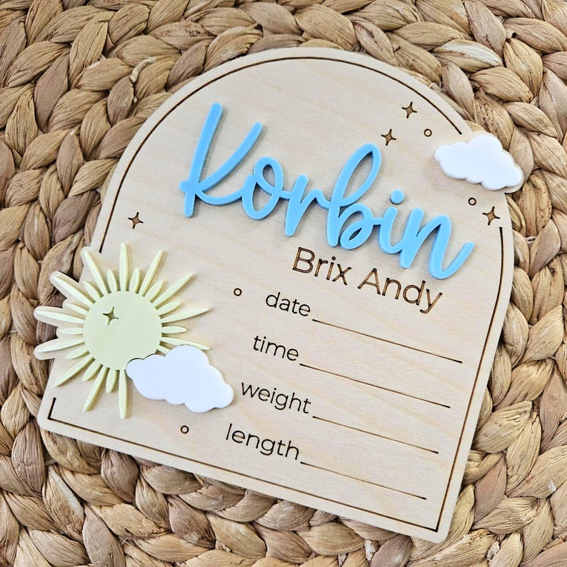 May include: An arched wooden birth announcement sign featuring the light blue 3D name Korbin, the name Brix Andy, and engraved lines for date, time, weight, and length. The sign includes a pale yellow sun, white clouds, and small star icons.