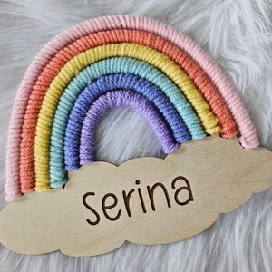 May include: A colorful rainbow-shaped wall hanging with a wooden cloud base. The rainbow features pastel shades of pink, orange, yellow, green, blue, and purple. The name "Serina" is engraved on the cloud. The item is set against a white, fluffy background.