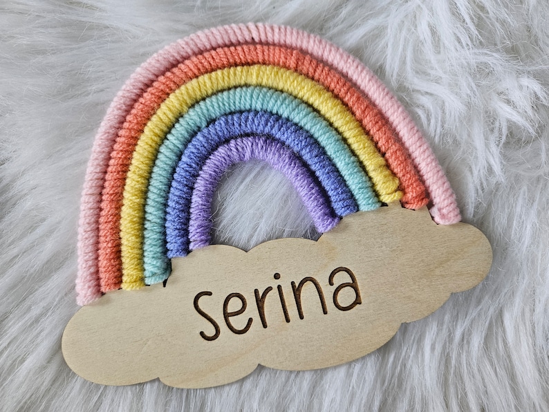 May include: A colorful rainbow-shaped wall hanging with a wooden cloud base. The rainbow features pastel shades of pink, orange, yellow, green, blue, and purple. The name "Serina" is engraved on the cloud. The item is set against a white, fluffy background.