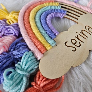 May include: A colorful rainbow wall hanging with a wooden cloud-shaped name tag that reads "Serina". The rainbow features shades of pink, orange, yellow, green, blue, and purple. Bundles of yarn in matching colors are arranged below.