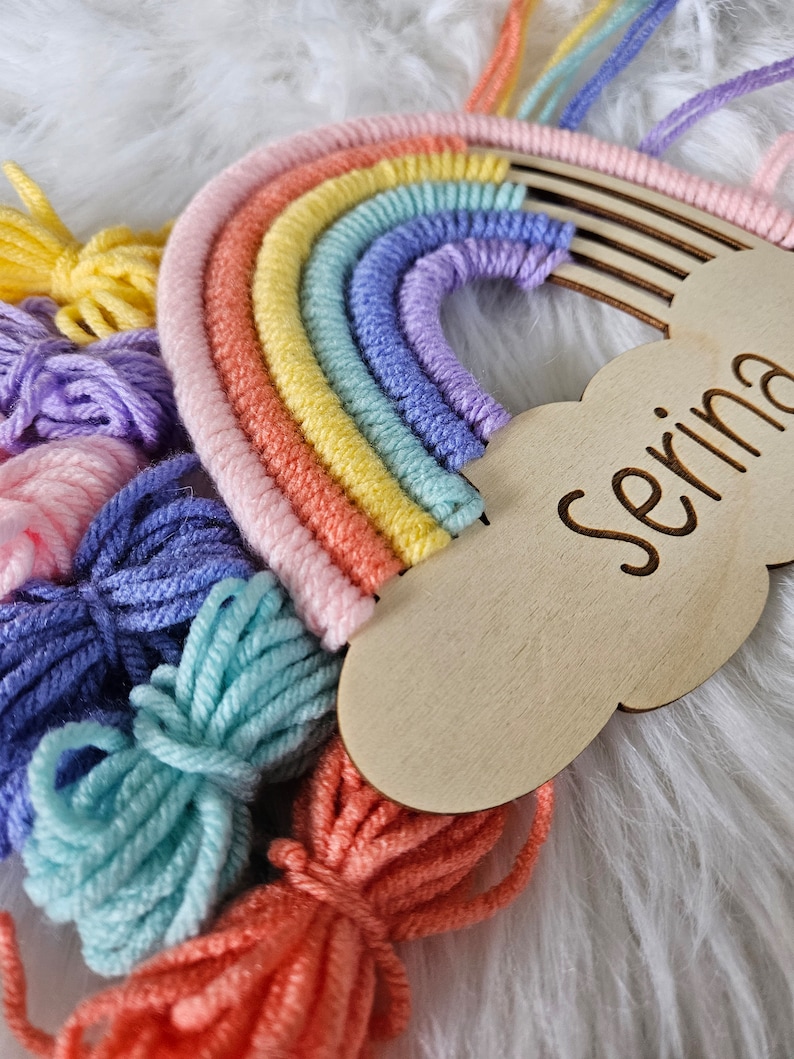 May include: A colorful rainbow wall hanging with a wooden cloud-shaped name tag that reads "Serina". The rainbow features shades of pink, orange, yellow, green, blue, and purple. Bundles of yarn in matching colors are arranged below.
