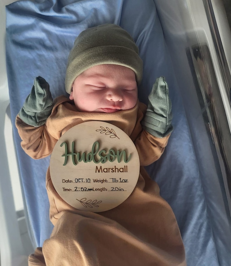 May include: A newborn wearing a tan long-sleeve gown, olive green beanie, and matching mittens. A round wooden plaque rests on the chest, engraved with the name Hudson Marshall, date OCT. 10, weight 7lb 1oz, time 2:52am, and length 20in.
