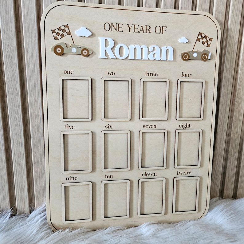 以下が含まれることがあります： 「ONE YEAR OF Roman」の文字が入った木製のマイルストーンボード。12個の写真スロット、レーシングカーと雲のアクセントがあります。1から12までの数字が各写真スロットの下に印刷されています。「Roman」という名前は白い3D文字です。