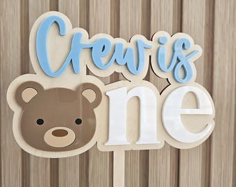 May include: A wooden cake topper featuring a light brown bear face, the text "Crew is" in light blue script, and the word "one" in white block letters. The design is layered on a light wood backing with a single stick for insertion into a cake.