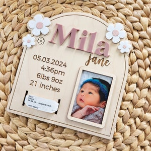 May include: A personalized wooden baby announcement plaque with the name "Mila" in pink letters. It includes the baby's birth date, time, weight (6lbs 9oz), and length (21 inches), along with a small photo and flower accents.