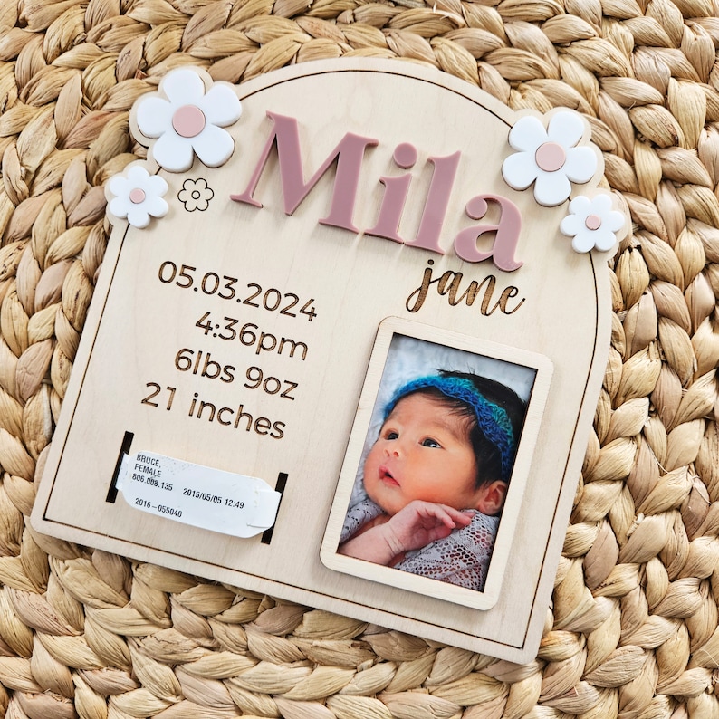 May include: A personalized wooden baby announcement plaque with the name "Mila" in pink letters. It includes the baby's birth date, time, weight (6lbs 9oz), and length (21 inches), along with a small photo and flower accents.