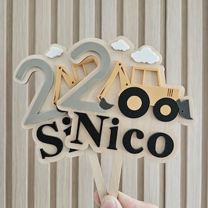 May include: A wooden cake topper featuring the number 2 in grey, a yellow construction excavator, white clouds, and the name Nico in black lettering. The design is layered on a light wood base with two sticks extending from the bottom for insertion.