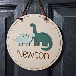 May include: A circular wooden sign with a light brown finish, featuring two dinosaur cutouts in shades of green and blue. The name "Newton" is laser-cut in a script font. A brown leather cord is used for hanging.