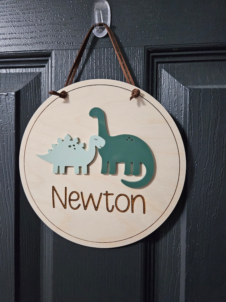 May include: A circular wooden sign with a light brown finish, featuring two dinosaur cutouts in shades of green and blue. The name "Newton" is laser-cut in a script font. A brown leather cord is used for hanging.