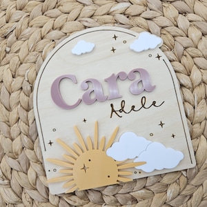 May include: An arched wooden plaque featuring the name Cara Adele in raised mauve lettering. The design includes white acrylic clouds, a yellow sun with radiating rays, and engraved star and circle accents on a light-colored wood surface.
