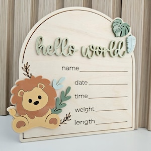 May include: An arched wooden birth announcement sign featuring the text hello world in raised sage green letters. The sign includes a cartoon lion figure, green leaf accents, and printed lines for name, date, time, weight, and length details.