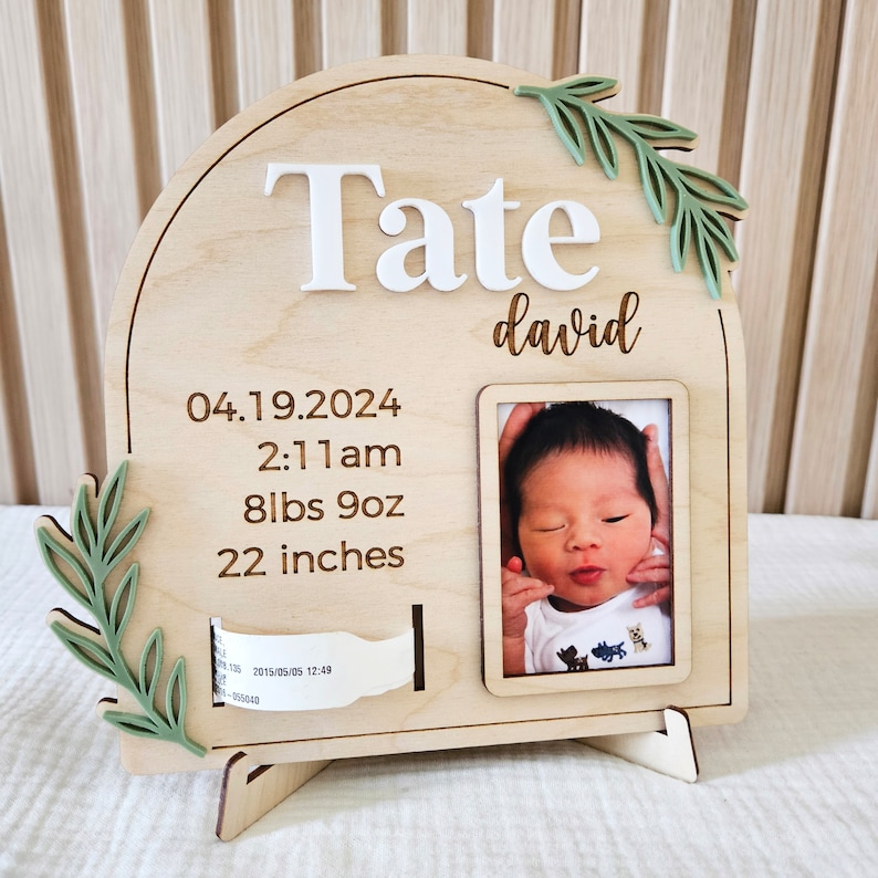 May include: A wooden baby announcement plaque with the name "Tate" in white and "david" in script. It includes the birth date, time, weight (8lbs 9oz), and length (22 inches). A photo frame holds a baby picture, and green leaf accents adorn the sides.