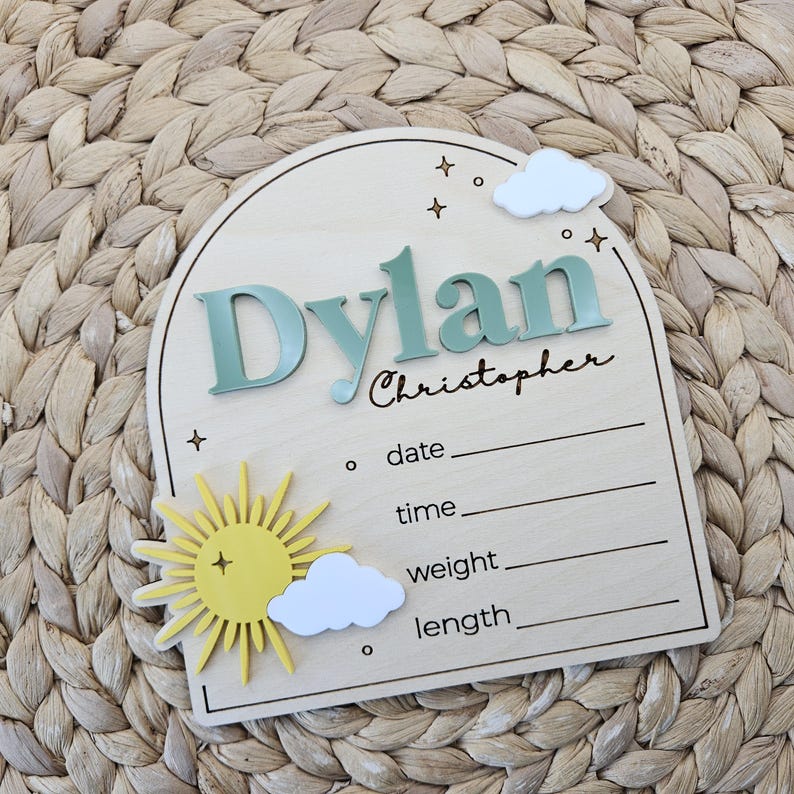 May include: An arched wooden birth announcement sign featuring the name Dylan Christopher in raised sage green and black script. The sign includes a yellow sun, white clouds, and engraved lines for date, time, weight, and length, accented with stars.