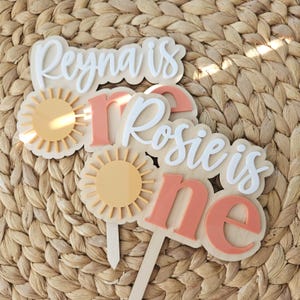 May include: Wooden cake toppers with the phrases "Reyna is one" and "Rosie is one". The toppers feature a sun design in yellow and peach, with white and peach lettering. The toppers are designed for a first birthday celebration.