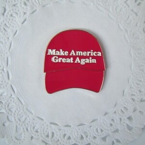 K&ouml;nnte beinhalten: Roter, hutformiger Ansteckpin mit dem wei&szlig;en Text "Make America Great Again". Der Pin befindet sich vor einem wei&szlig;en, spitzenartigen Hintergrund. Der Pin ist ein politisches Statement.