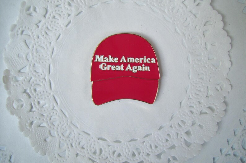 K&ouml;nnte beinhalten: Roter, hutformiger Ansteckpin mit dem wei&szlig;en Text "Make America Great Again". Der Pin befindet sich vor einem wei&szlig;en, spitzenartigen Hintergrund. Der Pin ist ein politisches Statement.