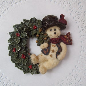 May include: A white snowman figurine wearing a brown hat and red scarf, holding a green holly wreath with red berries.