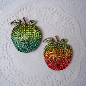 May include: Two apple-shaped brooches, one green and the other red, orange, and yellow, are adorned with sparkling rhinestones. Each brooch has a stem and two leaves. The brooches are displayed on a white, lacy doily.