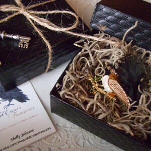 K&ouml;nnte beinhalten: Zwei schwarze Geschenkboxen, eine mit Bindfaden und einem silbernen Schl&uuml;ssel obenauf. Die andere Box ist ge&ouml;ffnet und zeigt eine Vogelbrosche, die in Stroh gebettet ist. Eine Visitenkarte mit einer Kr&auml;henillustration ist ebenfalls sichtbar.
