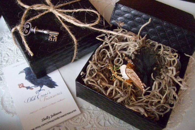 K&ouml;nnte beinhalten: Zwei schwarze Geschenkboxen, eine mit Bindfaden und einem silbernen Schl&uuml;ssel obenauf. Die andere Box ist ge&ouml;ffnet und zeigt eine Vogelbrosche, die in Stroh gebettet ist. Eine Visitenkarte mit einer Kr&auml;henillustration ist ebenfalls sichtbar.
