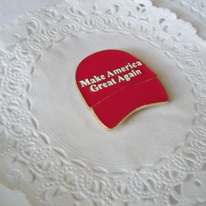 K&ouml;nnte beinhalten: Ein roter Emaille-Anstecker in Form einer Baseballkappe mit dem wei&szlig;en Text "Make America Great Again". Der Anstecker hat eine goldfarbene Umrandung und liegt auf einer wei&szlig;en, spitzenartigen Papierunterlage.
