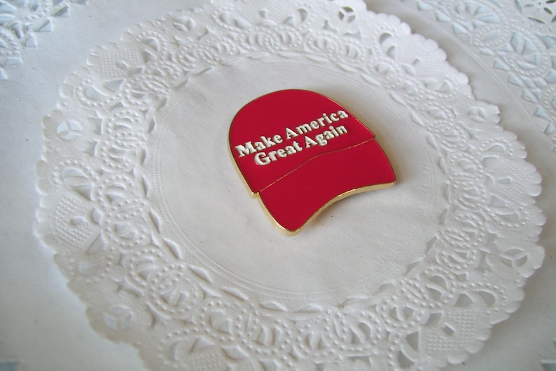 K&ouml;nnte beinhalten: Ein roter Emaille-Anstecker in Form einer Baseballkappe mit dem wei&szlig;en Text "Make America Great Again". Der Anstecker hat eine goldfarbene Umrandung und liegt auf einer wei&szlig;en, spitzenartigen Papierunterlage.
