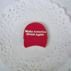 K&ouml;nnte beinhalten: Rote Baseballkappen-Anstecknadel mit wei&szlig;em Text "Make America Great Again". Die Anstecknadel befindet sich auf einem wei&szlig;en, spitzenartigen Hintergrund.