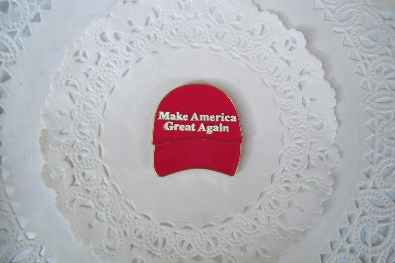 K&ouml;nnte beinhalten: Rote Baseballkappen-Anstecknadel mit wei&szlig;em Text "Make America Great Again". Die Anstecknadel befindet sich auf einem wei&szlig;en, spitzenartigen Hintergrund.
