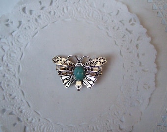 May include: A silver-toned butterfly brooch with intricate wing detailing and a central turquoise-colored stone. The brooch is set against a white, lacy background, creating a delicate and vintage aesthetic.