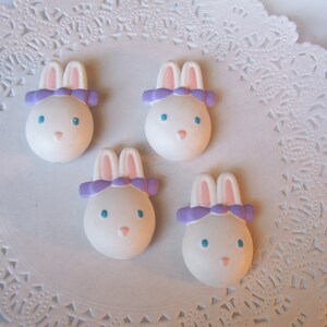 May include: Four white ceramic bunny figurines with purple bows. The bunnies are sitting on a white doily.