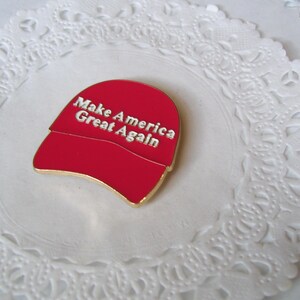 K&ouml;nnte beinhalten: Eine rote, hutformige Anstecknadel mit dem Text "Make America Great Again" in silbernen Buchstaben. Die Anstecknadel hat einen goldfarbenen Rand und liegt auf einer wei&szlig;en, spitzenartigen Unterlage.