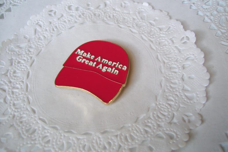 K&ouml;nnte beinhalten: Eine rote, hutformige Anstecknadel mit dem Text "Make America Great Again" in silbernen Buchstaben. Die Anstecknadel hat einen goldfarbenen Rand und liegt auf einer wei&szlig;en, spitzenartigen Unterlage.