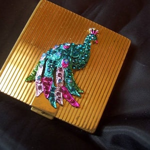 May include: A gold compact with a peacock design. The peacock is made of rhinestones and has a blue, green, and pink color scheme.