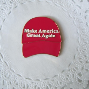K&ouml;nnte beinhalten: Roter Emaille-Anstecker in Form einer Baseballkappe mit dem wei&szlig;en Schriftzug "Make America Great Again". Der Anstecker liegt auf einem wei&szlig;en, spitzenartigen Hintergrund.
