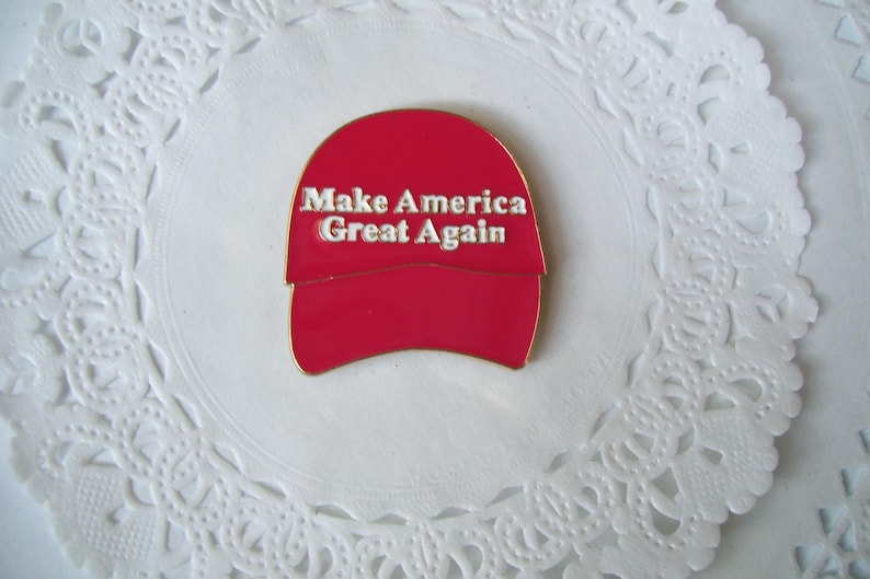 K&ouml;nnte beinhalten: Roter Emaille-Anstecker in Form einer Baseballkappe mit dem wei&szlig;en Schriftzug "Make America Great Again". Der Anstecker liegt auf einem wei&szlig;en, spitzenartigen Hintergrund.