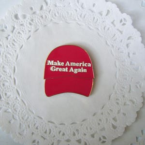 K&ouml;nnte beinhalten: Ein roter Emaille-Pin in Form einer Baseballkappe mit dem wei&szlig;en Schriftzug "Make America Great Again". Der Pin ist auf einer wei&szlig;en, spitzenartigen Papier-Serviette platziert.