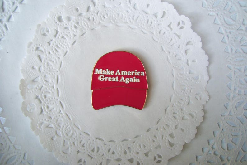 K&ouml;nnte beinhalten: Ein roter Emaille-Pin in Form einer Baseballkappe mit dem wei&szlig;en Schriftzug "Make America Great Again". Der Pin ist auf einer wei&szlig;en, spitzenartigen Papier-Serviette platziert.