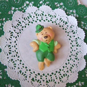 May include: A green and brown teddy bear wearing a green hat and a green vest. The bear is standing on a white doily with a green lace border.