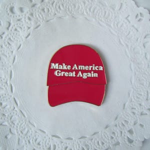K&ouml;nnte beinhalten: Rote, hutformige Anstecknadel mit dem wei&szlig;en Text "Make America Great Again". Die Anstecknadel ist auf einer wei&szlig;en, spitzenartigen Papier-Serviette zentriert. Die Anstecknadel hat einen goldenen Rand.