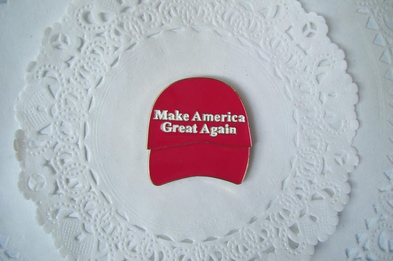 K&ouml;nnte beinhalten: Rote, hutformige Anstecknadel mit dem wei&szlig;en Text "Make America Great Again". Die Anstecknadel ist auf einer wei&szlig;en, spitzenartigen Papier-Serviette zentriert. Die Anstecknadel hat einen goldenen Rand.