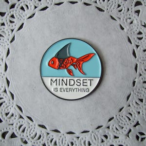 May include: A round enamel pin featuring a red fish with a shark fin, set against a light blue background. The pin has the text "MINDSET IS EVERYTHING" in white. The pin is on a white doily.