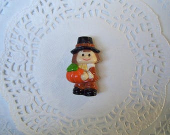 May include: A small plastic figurine of a person wearing a black pilgrim hat and brown clothing, holding a bright orange pumpkin with a green stem. The figure has brown hair, a painted face with a smile, and black shoes, standing in a front-facing pose.