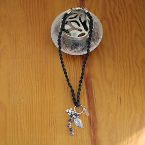 May include: A black cord necklace with a silver charm featuring a flower, leaves, and a crystal. The necklace is displayed on a wooden surface.
