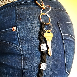 May include: A key chain with a key, a braided cord, and decorative beads. The key chain is attached to a pair of jeans.