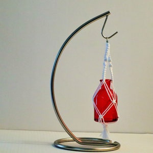 May include: A red glass bottle is suspended from a white macrame plant hanger. The hanger is attached to a silver metal stand.