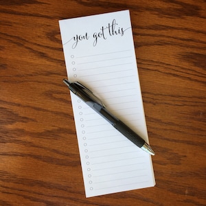 May include: A white notepad with a black pen lying on top. The notepad has a black and white inscription that says "you got this" and has a lined interior with a series of circles for check marks.
