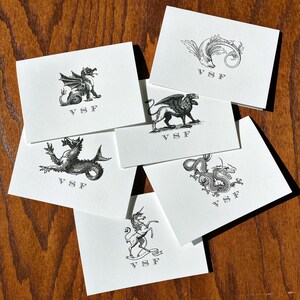 May include: A collection of white stationery cards featuring various mythical creatures and decorative elements in black ink. Each card displays the initials "VSF". The cards are arranged on a wooden surface.