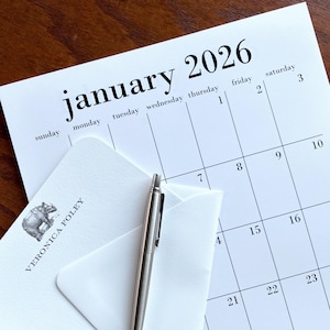 May include: A close-up of a January 2026 calendar with a white envelope and a silver pen. The calendar is open to the first week of the month. The envelope has a bear illustration and the text "VERONICA FOLEY".