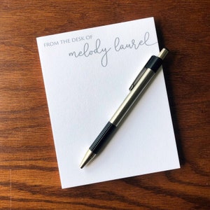 May include: A white notepad with the text "From the desk of melody laurel" printed in gray. A silver pen with a black grip is lying on top of the notepad.